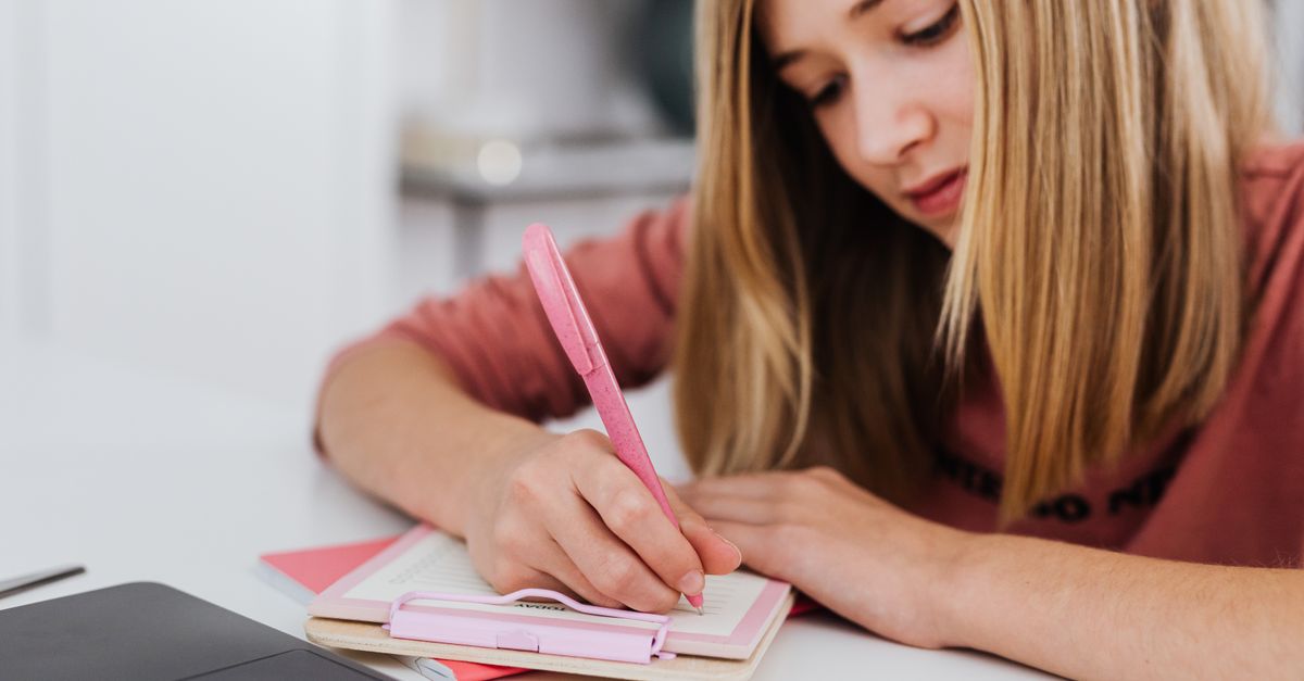 A focused young woman writing in a notebook indoors, showcasing creativity and study habits.